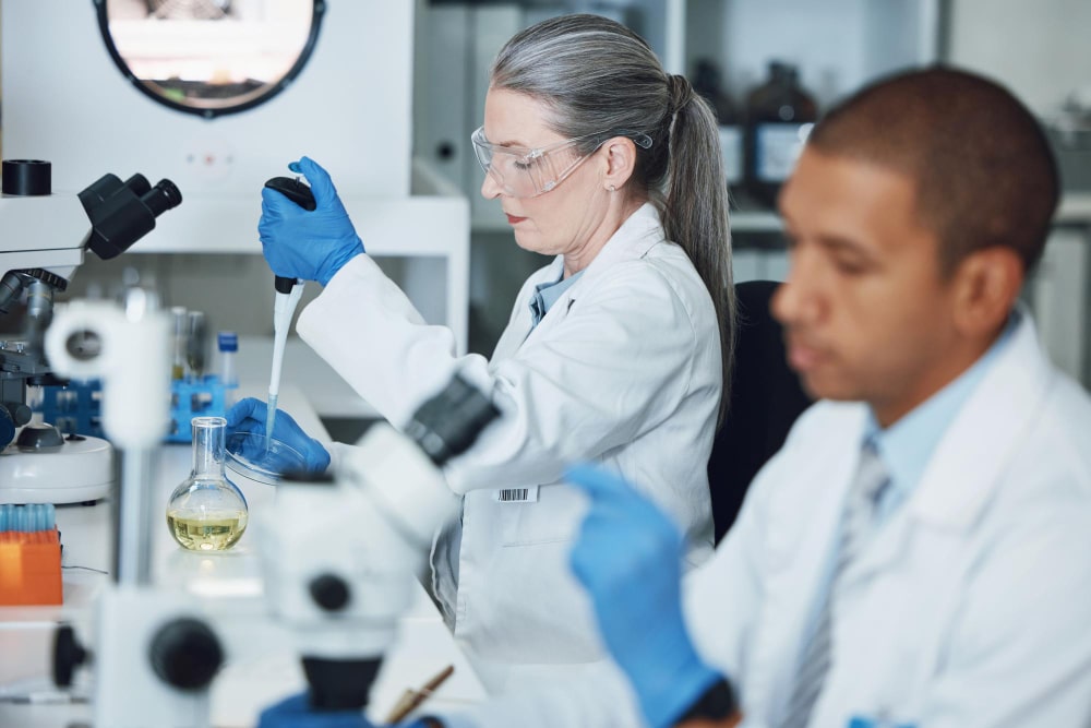 Researchers in lab coats and gloves performing experiments with microscopes and flasks in a biotechnology research laboratory.