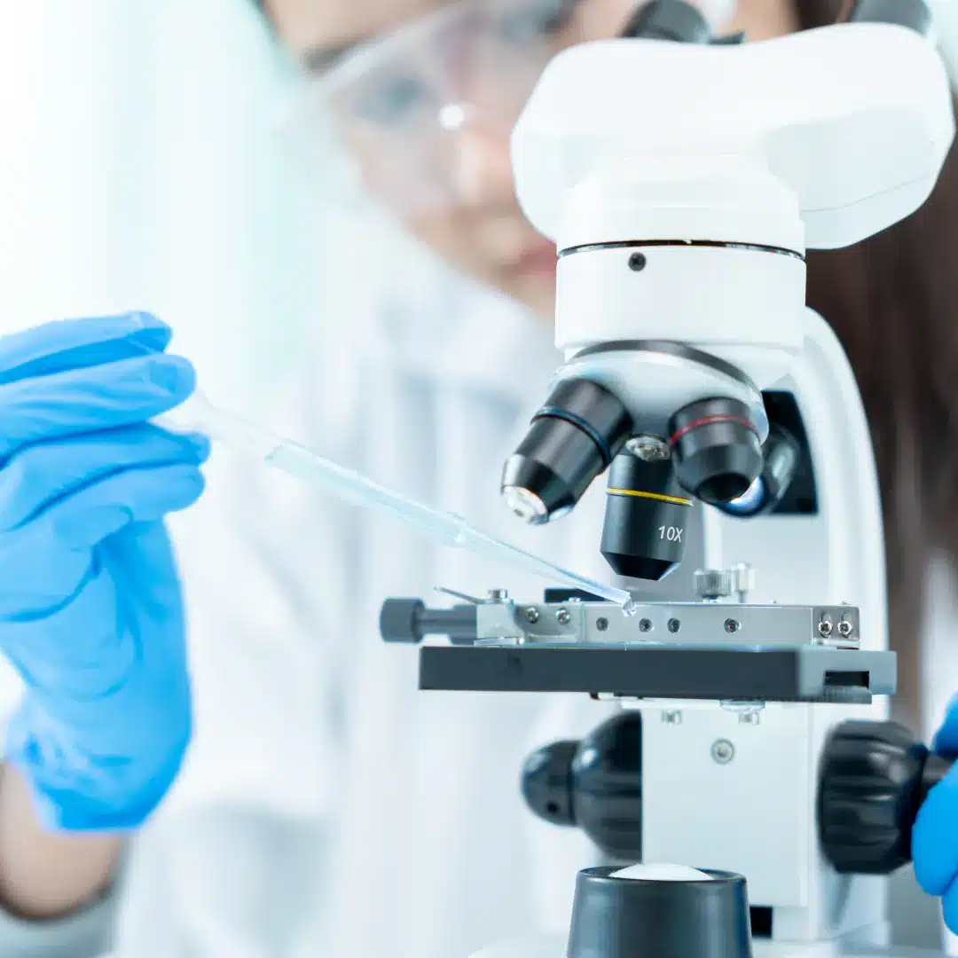 a person in a lab coat and gloves holding a pipette over a microscope