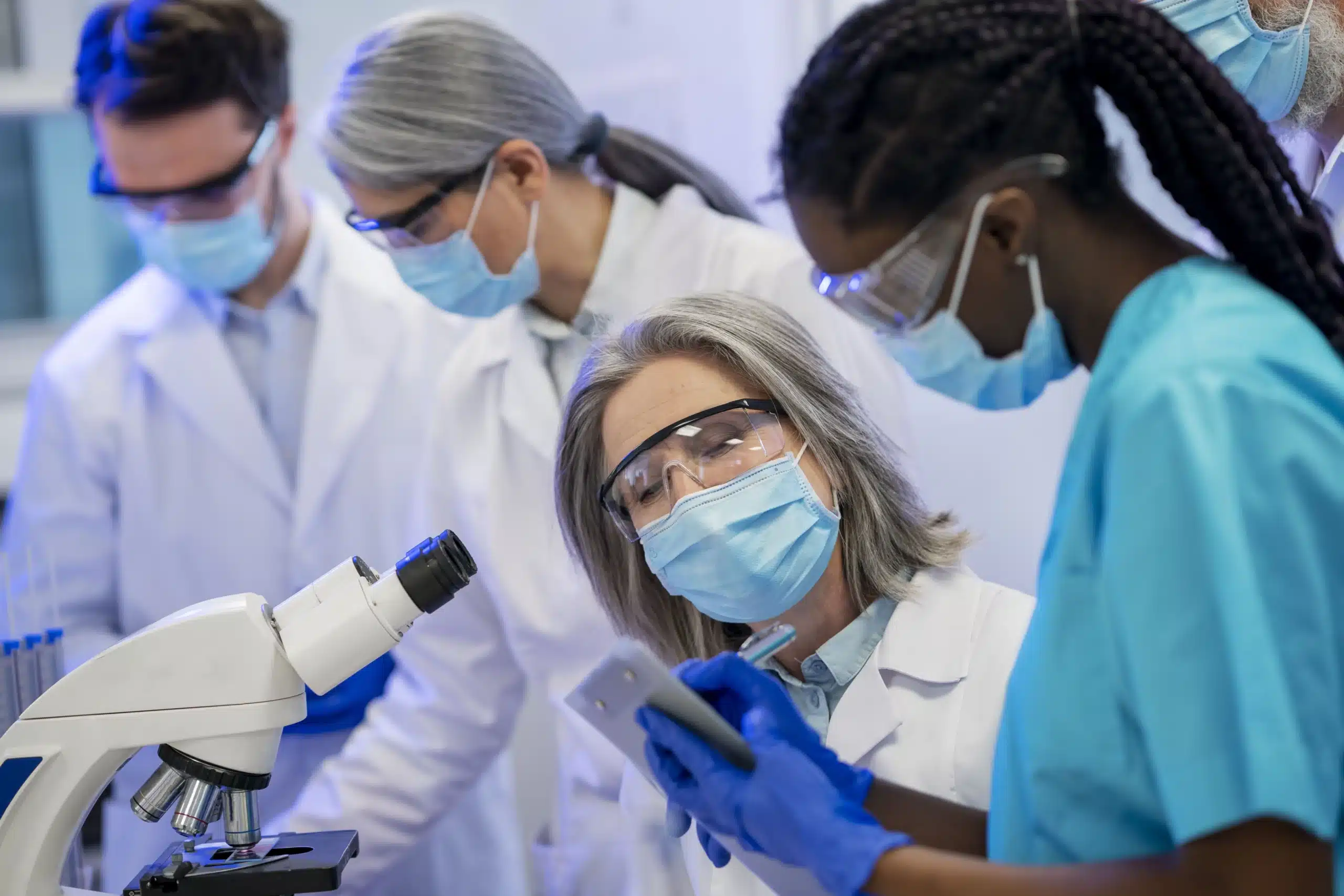 Group of scientists and medical researchers wearing masks and lab coats collaborating on cancer research using microscope and lab equipment.